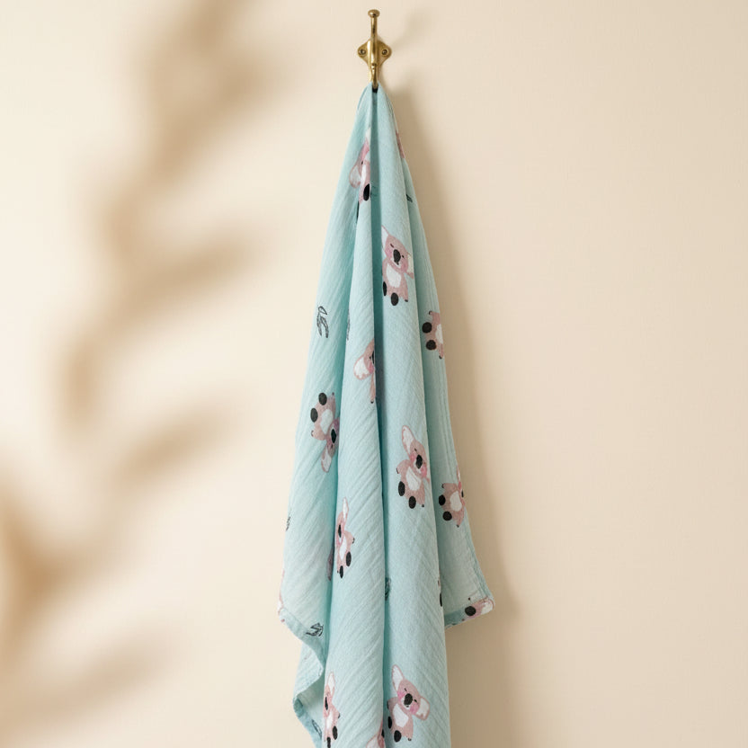 Premium mint teal muslin swaddle blanket with an adorable koala bear pattern hanging from an elegant brass wall hook by Little Babybox