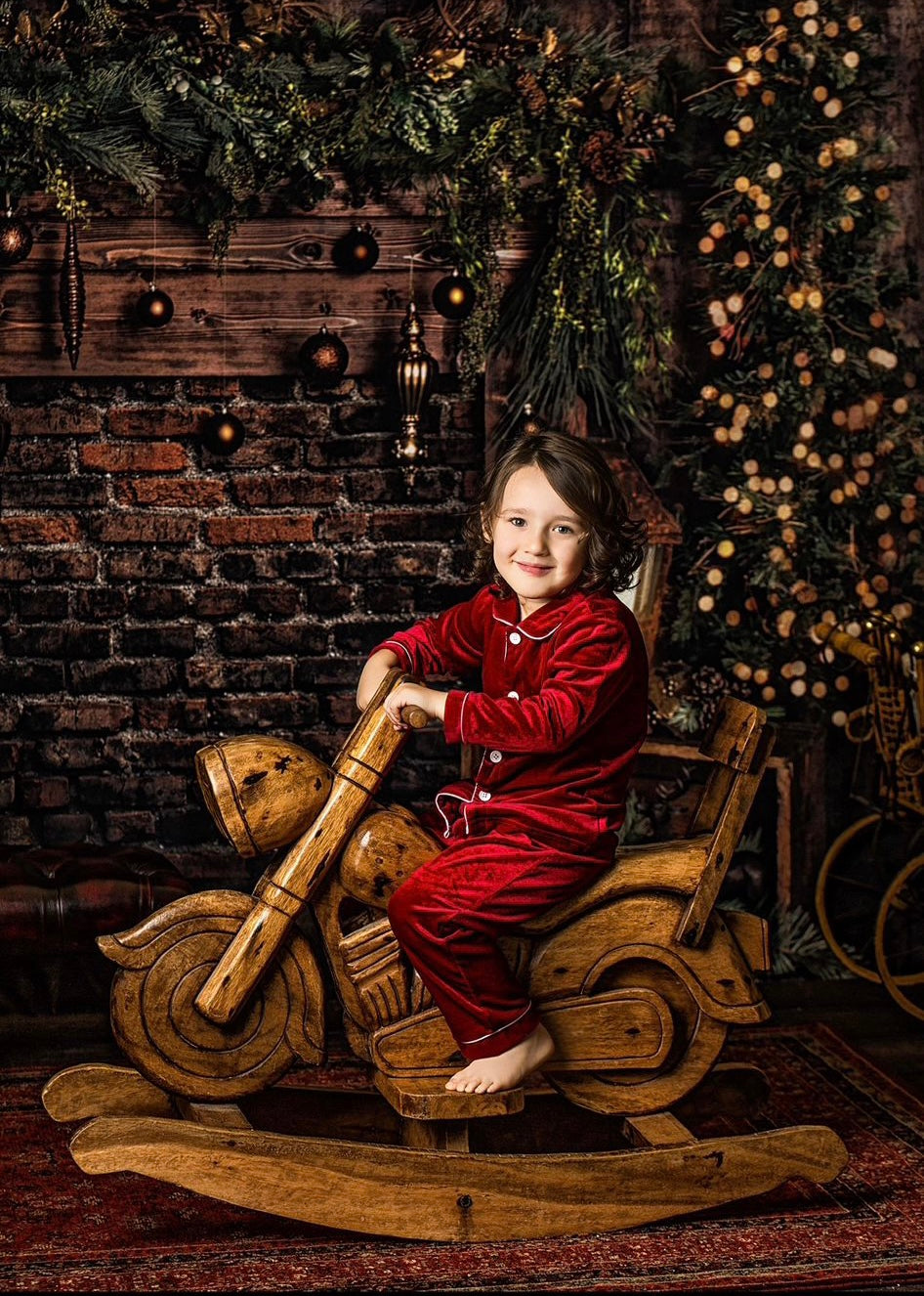 Child wearing red velvet Christmas pyjamas with white piping on a wooden rocking motorbike