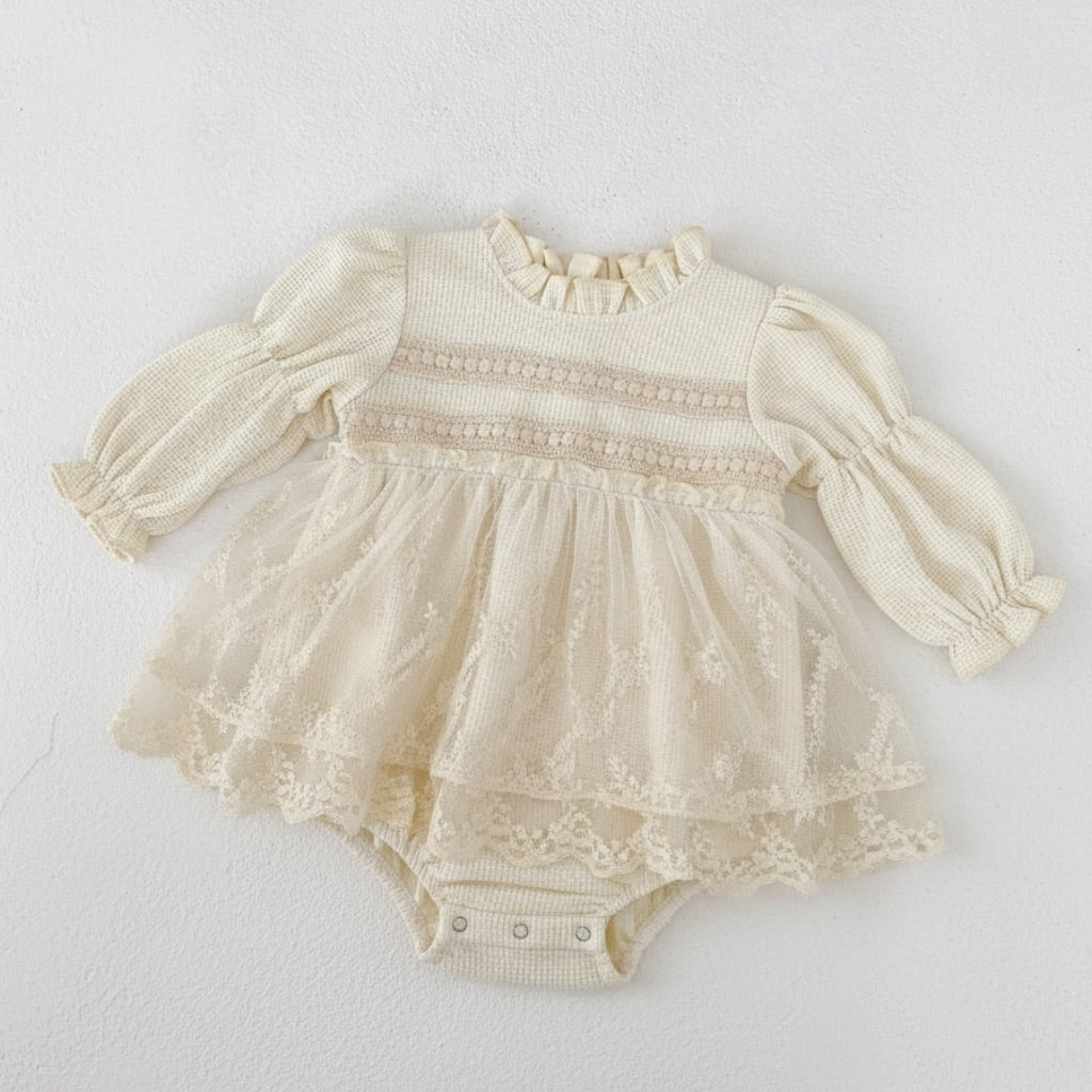 Oh Delilah long-sleeve romper featuring a waffle-textured knit bodice with a ruffled collar. The midsection is adorned with intricate beige lace inserts, flowing into a layered, sheer tulle and lace skirt overlay over a classic snap-closure bodysuit bottom.