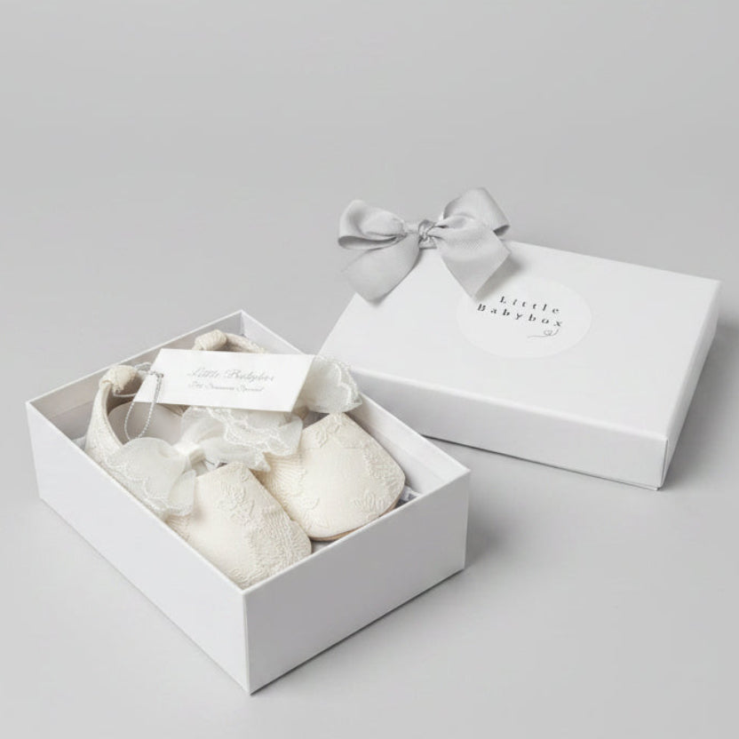 The Aurelia white lace fabric baby shoes neatly displayed inside their open white gift box next to the closed presentation box lid featuring a silver grey grosgrain ribbon bow