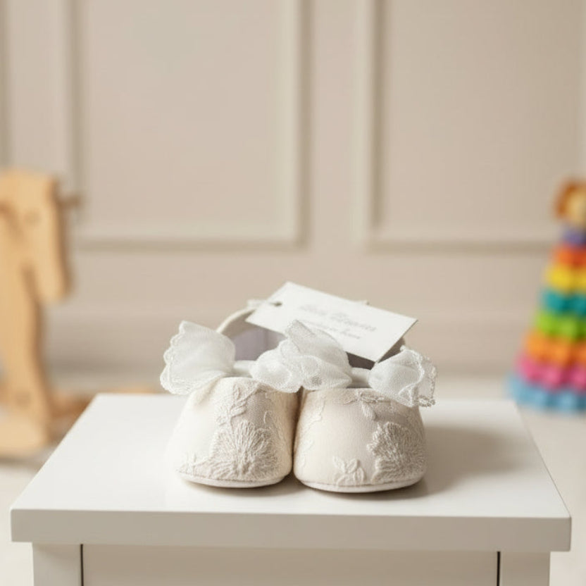 Pair of white Aurelia fabric baby shoes with prominent lace tulle bows sitting on a white wooden side table in a nursery background