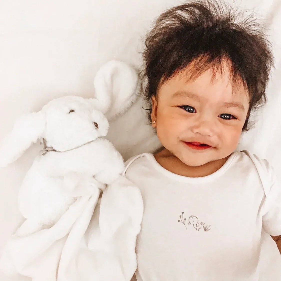 Cute smiling baby boy lying down in a white vest next to a luxury soft white BunnyBoo plush bunny comforter blanket by Little Babybox