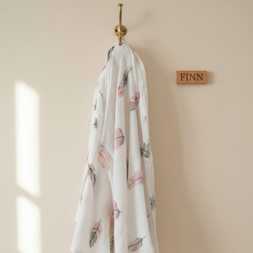 Luxury soft pastel colour feathers patterned premium muslin swaddle blanket hanging on a brass hook next to a personalised wooden name sign by Little Babybox