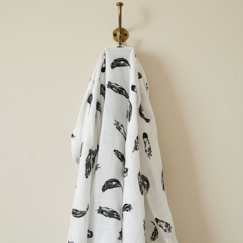 Luxury monochrome black and white feathers patterned premium muslin swaddle blanket hanging elegantly from a brass wall hook by Little Babybox