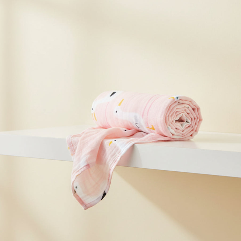 Luxury rolled pastel pink muslin swaddle blanket with a cute white duck pattern resting elegantly on a minimalist white shelf by Little Babybox