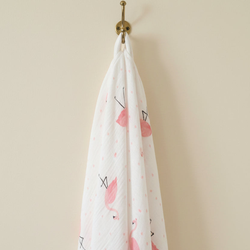 Premium white muslin swaddle blanket with a soft pink flamingo pattern hanging elegantly from a vintage brass wall hook by Little Babybox