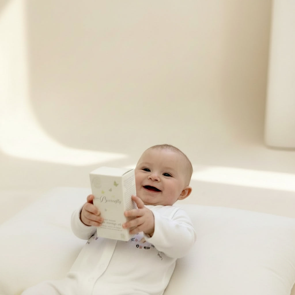 Happy newborn baby resting on a cream cushion while holding a Little Butterfly London product box from a luxury gift set.