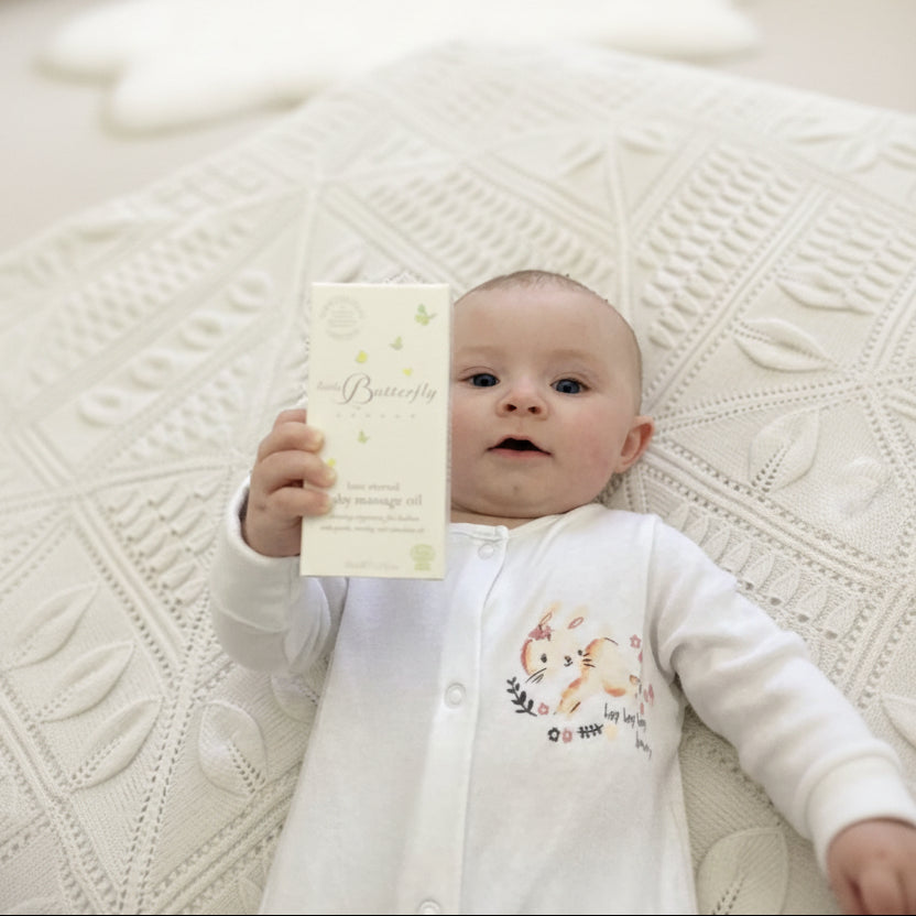 Baby lying on a knitted cream blanket holding a box of Little Butterfly London organic baby massage oil.